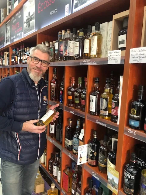 Laurent sourit devant le mur de whiskies de sa boutique, une bouteille à la main